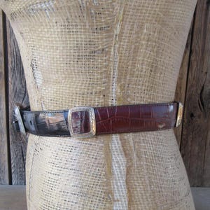 Pu&ograve; includere: Una cintura bicolore con fibbia e dettagli argentati. La cintura &egrave; realizzata in finta pelle di coccodrillo nera e marrone. La fibbia &egrave; quadrata con un design in rilievo. La cintura &egrave; esposta su un manichino.