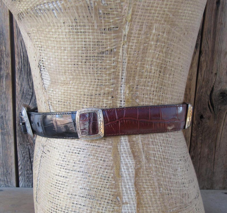 Pu&ograve; includere: Una cintura bicolore con fibbia e dettagli argentati. La cintura &egrave; realizzata in finta pelle di coccodrillo nera e marrone. La fibbia &egrave; quadrata con un design in rilievo. La cintura &egrave; esposta su un manichino.