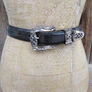 May include: A black belt with a silver buckle and clasp. The belt has a textured finish, and the buckle features an ornate design. The belt is displayed on a mannequin.