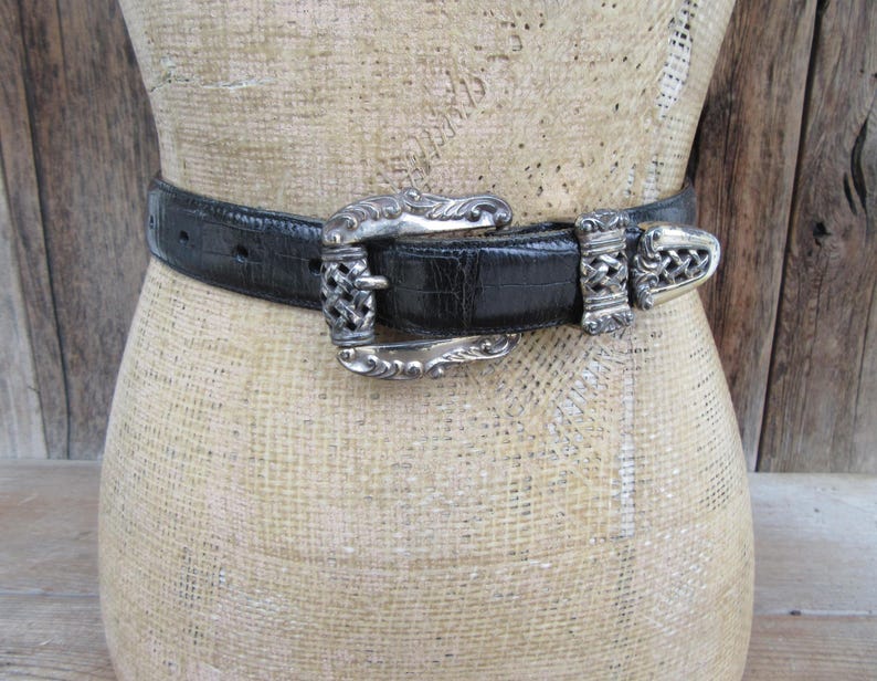 May include: A black belt with a silver buckle and clasp. The belt has a textured finish, and the buckle features an ornate design. The belt is displayed on a mannequin.