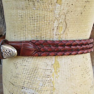 Op de afbeelding: Een bruine leren riem met een gevlochten ontwerp en een zilveren gesp. De riem is om een ​​mannequin gewikkeld. De gesp heeft een sierlijk ontwerp. De riem heeft een bruine kleur.