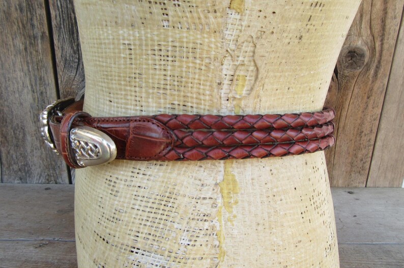 Op de afbeelding: Een bruine leren riem met een gevlochten ontwerp en een zilveren gesp. De riem is om een ​​mannequin gewikkeld. De gesp heeft een sierlijk ontwerp. De riem heeft een bruine kleur.