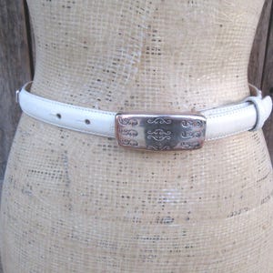 May include: A white leather belt with a decorative silver buckle. The buckle has an ornate design. The belt is shown on a mannequin. The belt is approximately 1 inch wide.
