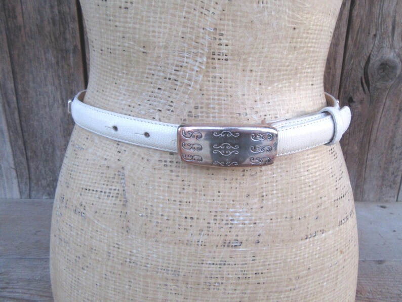 May include: A white leather belt with a decorative silver buckle. The buckle has an ornate design. The belt is shown on a mannequin. The belt is approximately 1 inch wide.