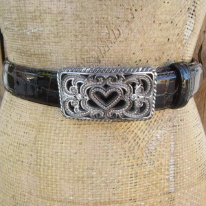 Op de afbeelding: Een zwarte riem met een zilveren gesp met een hartvormig ontwerp. De riem heeft een textuur met krokodillenpatroon. De gesp is versierd met kleine, sprankelende versieringen, die een vleugje elegantie aan het accessoire toevoegen.