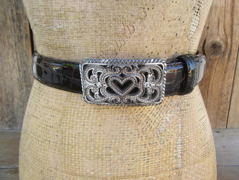 Op de afbeelding: Een zwarte riem met een zilveren gesp met een hartvormig ontwerp. De riem heeft een textuur met krokodillenpatroon. De gesp is versierd met kleine, sprankelende versieringen, die een vleugje elegantie aan het accessoire toevoegen.