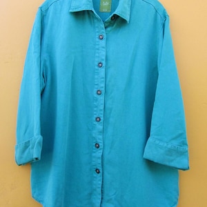 May include: A turquoise button-down shirt with long sleeves, cuffed at the elbows. The shirt has a classic collar and a curved hem. The shirt is hanging on a hanger against a yellow background. The shirt has a small label near the collar.