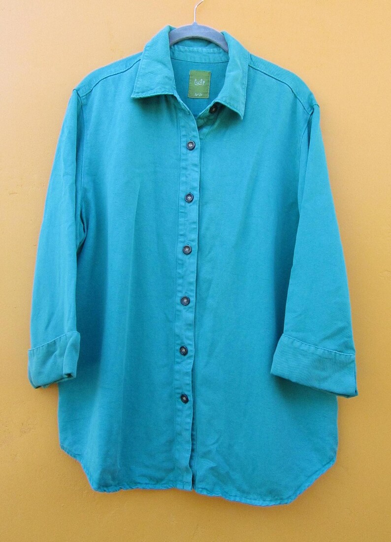 May include: A turquoise button-down shirt with long sleeves, cuffed at the elbows. The shirt has a classic collar and a curved hem. The shirt is hanging on a hanger against a yellow background. The shirt has a small label near the collar.