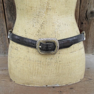 K&ouml;nnte beinhalten: Ein schwarzer G&uuml;rtel mit strukturiertem Muster und einer silber- und goldfarbenen Schnalle. Der G&uuml;rtel wird auf einer Schaufensterpuppe pr&auml;sentiert. Die Schnalle hat eine rechteckige Form mit dekorativem Design.