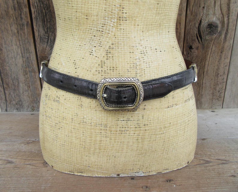 K&ouml;nnte beinhalten: Ein schwarzer G&uuml;rtel mit strukturiertem Muster und einer silber- und goldfarbenen Schnalle. Der G&uuml;rtel wird auf einer Schaufensterpuppe pr&auml;sentiert. Die Schnalle hat eine rechteckige Form mit dekorativem Design.