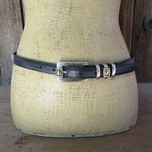 May include: Black leather belt with a silver buckle and two silver loops with gold-colored accents. The belt is displayed on a mannequin with a wooden background.