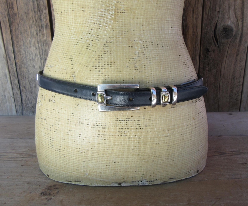 May include: Black leather belt with a silver buckle and two silver loops with gold-colored accents. The belt is displayed on a mannequin with a wooden background.