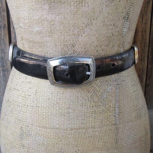 Pu&ograve; includere: Una cintura nera con fibbia e dettagli argentati. La cintura ha una superficie strutturata ed &egrave; allacciata attorno a un manichino. La fibbia &egrave; rettangolare con un design ornato.