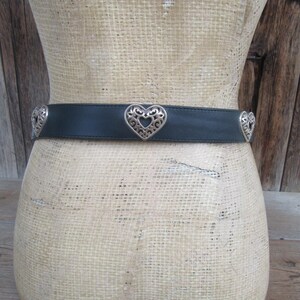 Op de afbeelding: Een zwarte leren riem met zilveren hartvormige versieringen. De riem wordt getoond op een mannequin. De hartvormige accenten hebben openwerk ontwerpen. De achtergrond is een houten muur.