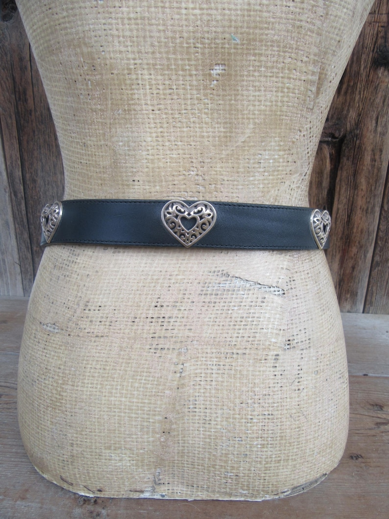 Op de afbeelding: Een zwarte leren riem met zilveren hartvormige versieringen. De riem wordt getoond op een mannequin. De hartvormige accenten hebben openwerk ontwerpen. De achtergrond is een houten muur.