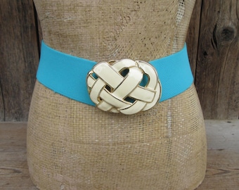 May include: A turquoise elastic belt with a cream-colored, gold-trimmed knot-shaped buckle. The belt is displayed on a mannequin. The background is a wooden surface.