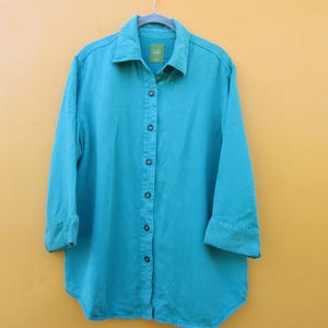 May include: A turquoise button-down shirt with long sleeves and a classic collar. The shirt is hanging on a metal hanger against a yellow background. The sleeves are cuffed at the elbows.