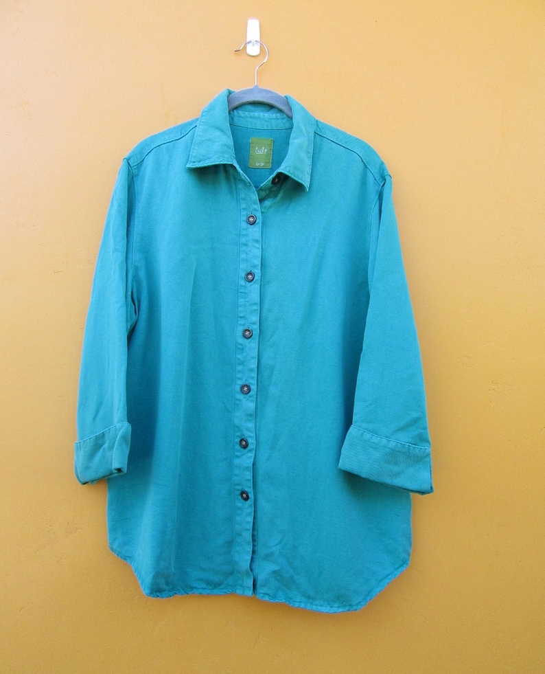May include: A turquoise button-down shirt with long sleeves and a classic collar. The shirt is hanging on a metal hanger against a yellow background. The sleeves are cuffed at the elbows.