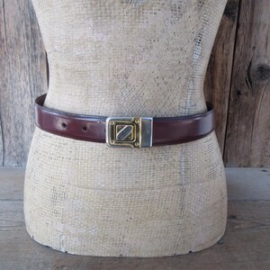 May include: A brown leather belt with a gold and silver buckle is displayed on a mannequin. The belt is approximately 1.25 inches wide. The buckle has a rectangular shape with a gold-colored center and silver-colored edges. The belt is fastened around the mannequin's waist.