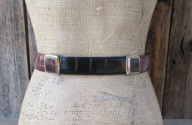 Pu&ograve; includere: Una cintura in pelle con una sezione centrale nera e estremit&agrave; bordeaux. La cintura ha una fibbia rettangolare argentata su ciascuna estremit&agrave;. La cintura &egrave; esposta su un manichino.