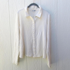 May include: A long-sleeved, off-white button-down shirt with a textured, crinkled fabric surface. The garment features a classic collar, a full-length front button placket with matching buttons, and straight cuffs, hanging on a dark gray hanger.