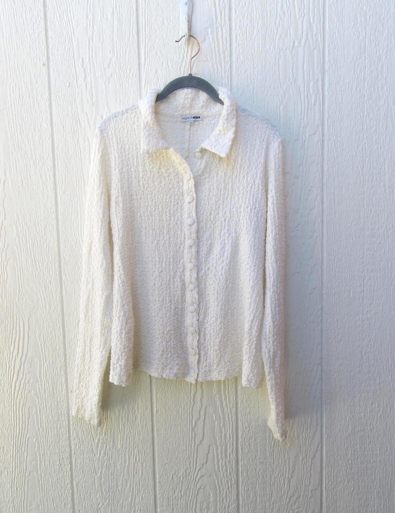 May include: A long-sleeved, off-white button-down shirt with a textured, crinkled fabric surface. The garment features a classic collar, a full-length front button placket with matching buttons, and straight cuffs, hanging on a dark gray hanger.