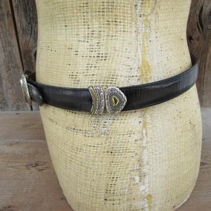 K&ouml;nnte beinhalten: Schwarzer Lederg&uuml;rtel mit silber- und goldfarbener Schnalle. Der G&uuml;rtel ist um eine Schaufensterpuppe gewickelt. Die Schnalle hat ein geometrisches Design mit einem dreieckigen Mittelteil. Der G&uuml;rtel ist strukturiert und scheint aus Leder zu sein.