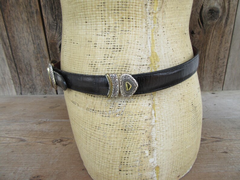 K&ouml;nnte beinhalten: Schwarzer Lederg&uuml;rtel mit silber- und goldfarbener Schnalle. Der G&uuml;rtel ist um eine Schaufensterpuppe gewickelt. Die Schnalle hat ein geometrisches Design mit einem dreieckigen Mittelteil. Der G&uuml;rtel ist strukturiert und scheint aus Leder zu sein.