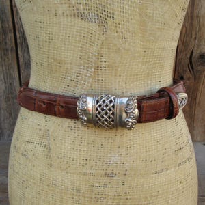 Op de afbeelding: Een bruine leren riem met een zilverkleurige gesp. De gesp heeft een geweven middenontwerp en sierlijke details aan de zijkanten. De riem wordt op een mannequin getoond.