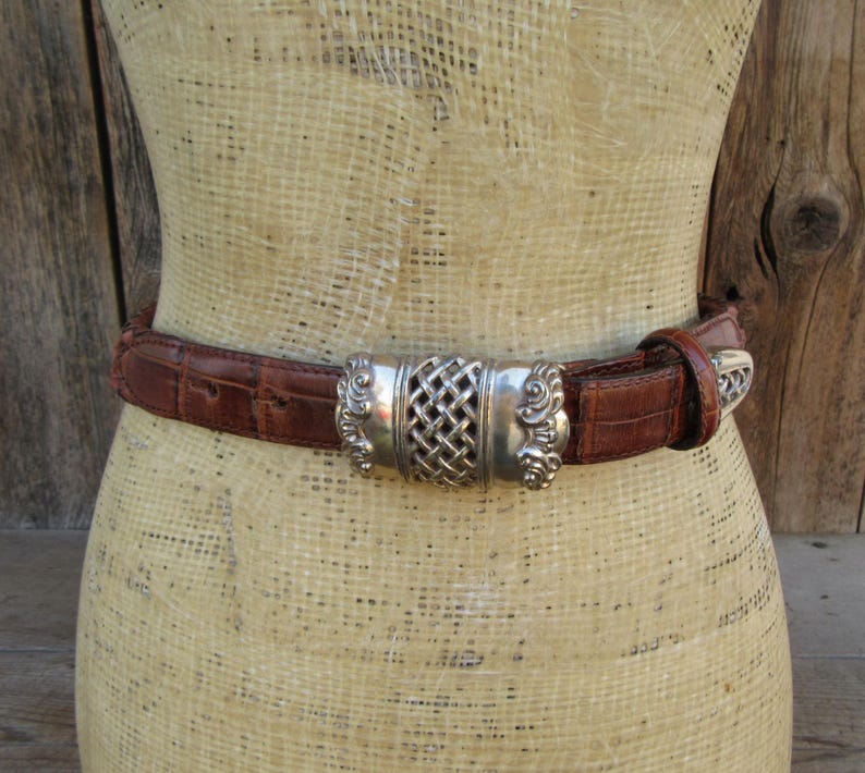 Op de afbeelding: Een bruine leren riem met een zilverkleurige gesp. De gesp heeft een geweven middenontwerp en sierlijke details aan de zijkanten. De riem wordt op een mannequin getoond.