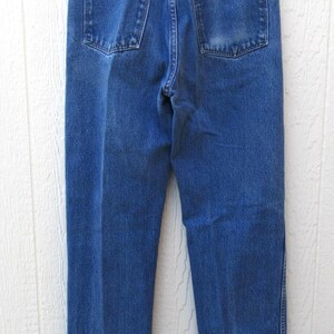 May include: Back view of blue denim jeans with two back pockets and a brown label. The jeans are hanging on a clear hanger against a white wooden background. The jeans have a classic, straight-leg fit.