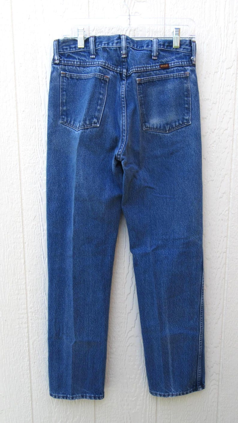 May include: Back view of blue denim jeans with two back pockets and a brown label. The jeans are hanging on a clear hanger against a white wooden background. The jeans have a classic, straight-leg fit.