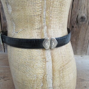 May include: Black belt with a silver buckle, displayed on a mannequin. The belt has a textured, crocodile-like pattern. The buckle is a decorative silver design with an ornate pattern.