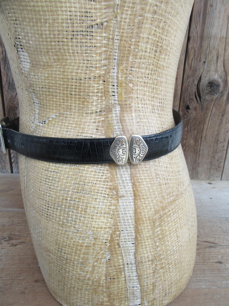 May include: Black belt with a silver buckle, displayed on a mannequin. The belt has a textured, crocodile-like pattern. The buckle is a decorative silver design with an ornate pattern.