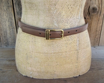 May include: A brown leather belt with a gold-colored buckle. The belt is displayed on a mannequin. The belt has a rectangular buckle with a small logo. The belt has several holes for adjustment.