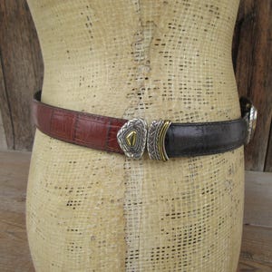 K&ouml;nnte beinhalten: Ein zweifarbiges G&uuml;rtel mit dekorativer Schnalle. Der G&uuml;rtel besteht aus braunem und schwarzem Leder. Die Schnalle ist silberfarben mit goldenen Akzenten. Der G&uuml;rtel wird auf einer Schaufensterpuppe pr&auml;sentiert.