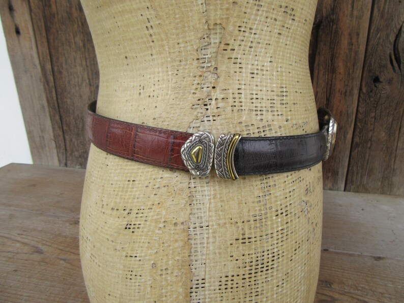 K&ouml;nnte beinhalten: Ein zweifarbiges G&uuml;rtel mit dekorativer Schnalle. Der G&uuml;rtel besteht aus braunem und schwarzem Leder. Die Schnalle ist silberfarben mit goldenen Akzenten. Der G&uuml;rtel wird auf einer Schaufensterpuppe pr&auml;sentiert.