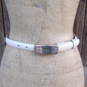 May include: A white leather belt with a silver rectangular buckle. The buckle has an ornate design. The belt is displayed on a mannequin. The background is a wooden wall.
