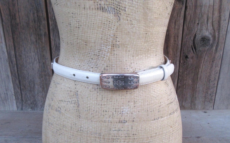 May include: A white leather belt with a silver rectangular buckle. The buckle has an ornate design. The belt is displayed on a mannequin. The background is a wooden wall.