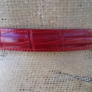 Puede incluir: Primer plano de un cintur&oacute;n de cuero rojo con un patr&oacute;n de cocodrilo. El cintur&oacute;n es de un tono rojo vibrante y presenta costuras visibles en los bordes. La textura del cuero es claramente visible, mostrando el dise&ntilde;o en relieve.