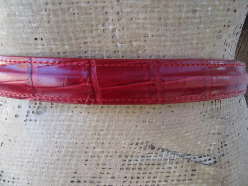 Puede incluir: Primer plano de un cintur&oacute;n de cuero rojo con un patr&oacute;n de cocodrilo. El cintur&oacute;n es de un tono rojo vibrante y presenta costuras visibles en los bordes. La textura del cuero es claramente visible, mostrando el dise&ntilde;o en relieve.