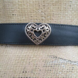 Op de afbeelding: Een zwarte leren riem met een zilveren hartvormige gesp. De gesp heeft een opengewerkt ontwerp. De riem wordt op een mannequin getoond.