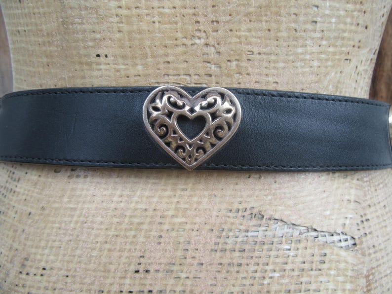 Op de afbeelding: Een zwarte leren riem met een zilveren hartvormige gesp. De gesp heeft een opengewerkt ontwerp. De riem wordt op een mannequin getoond.