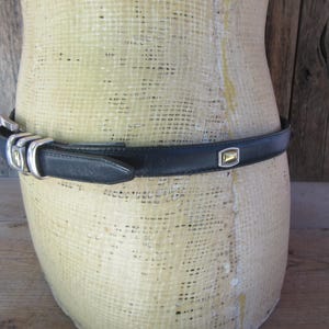May include: A black leather belt with a silver buckle and a gold-toned square accent. The belt is displayed on a mannequin. The belt is approximately 1 inch wide.