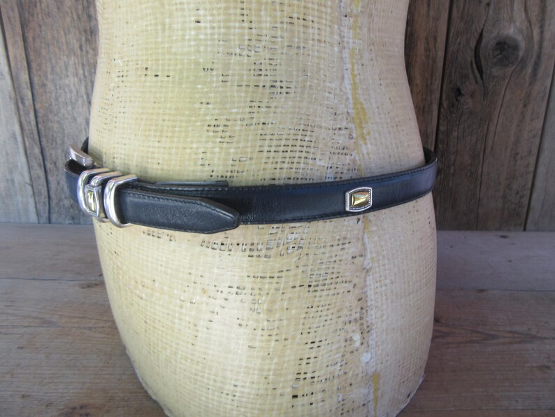 May include: A black leather belt with a silver buckle and a gold-toned square accent. The belt is displayed on a mannequin. The belt is approximately 1 inch wide.