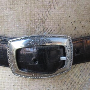 Pu&ograve; includere: Una cintura nera con fibbia e dettagli argentati. La cintura presenta un motivo strutturato a forma di coccodrillo. La fibbia argentata &egrave; rettangolare con un design ornato. La cintura &egrave; esposta su un manichino.