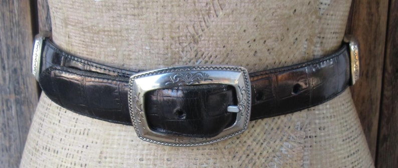 Pu&ograve; includere: Una cintura nera con fibbia e dettagli argentati. La cintura presenta un motivo strutturato a forma di coccodrillo. La fibbia argentata &egrave; rettangolare con un design ornato. La cintura &egrave; esposta su un manichino.