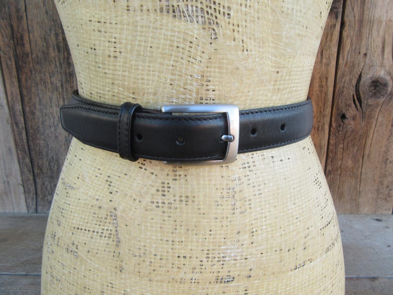 Pode incluir: Um cinto de couro preto com uma fivela retangular prateada. O cinto &eacute; usado em volta de um manequim. O cinto tem v&aacute;rios orif&iacute;cios para ajuste. O cinto tem cerca de 3,8 cm de largura.