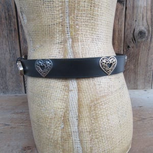 Op de afbeelding: Zwarte leren riem met zilveren hartvormige versieringen. De riem heeft een zilveren gesp en decoratieve zilveren accenten. De hartvormige accenten hebben een open filigraan ontwerp. De riem wordt getoond op een mannequin.