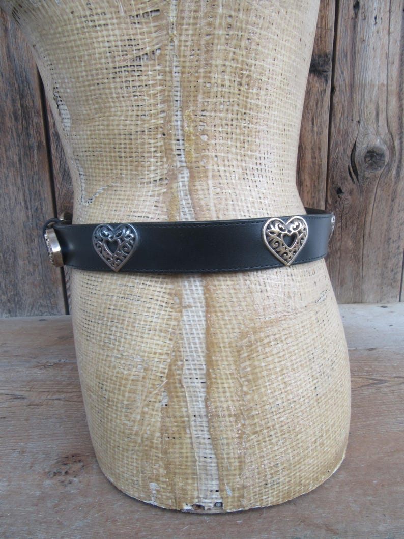 Op de afbeelding: Zwarte leren riem met zilveren hartvormige versieringen. De riem heeft een zilveren gesp en decoratieve zilveren accenten. De hartvormige accenten hebben een open filigraan ontwerp. De riem wordt getoond op een mannequin.